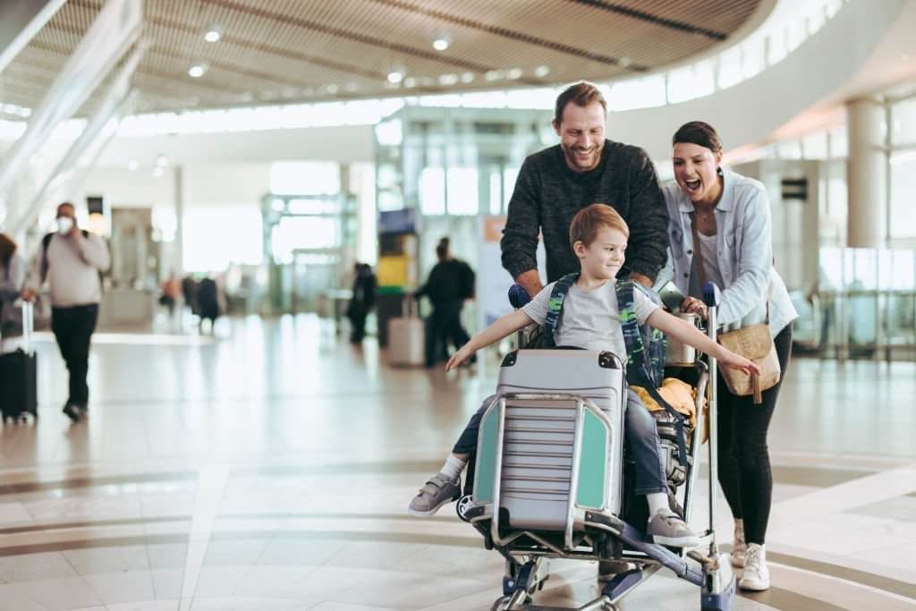 Parents pushing a luggage trolley with child sitting on it in airport on their family trip