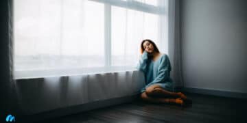 Woman in sky-blue top sitting on floor besides a curtained window-why attention fatigue is the result of too much stimulation