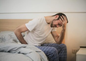 Middle aged man in white crew neck shirt and pajamas sitting on his bed in the morning feeling groggy holding his head