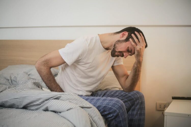 Middle aged man in white crew neck shirt and pajamas sitting on his bed in the morning feeling groggy holding his head