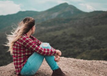 Young blonde women sitting on a hill in red check shirt and blue denim pants with wind blowing through her hair-i am broken
