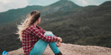 Young blonde women sitting on a hill in red check shirt and blue denim pants with wind blowing through her hair-i am broken
